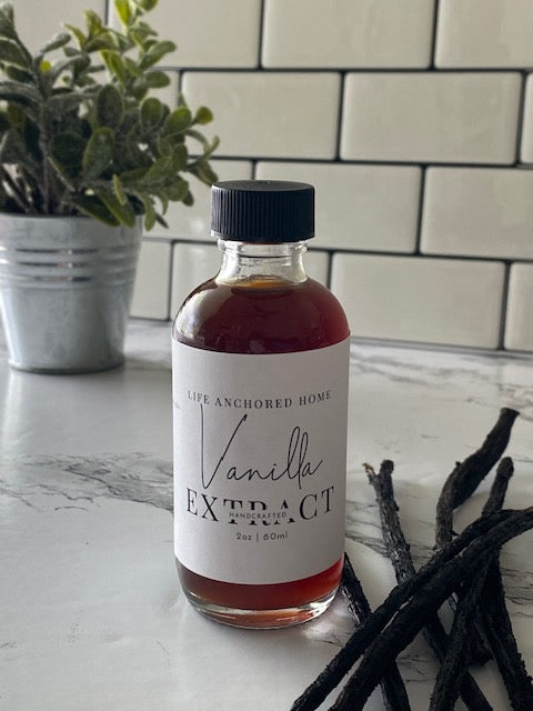 Bottle of vanilla extract on a marble surface with vanilla beans and a plant in the background.